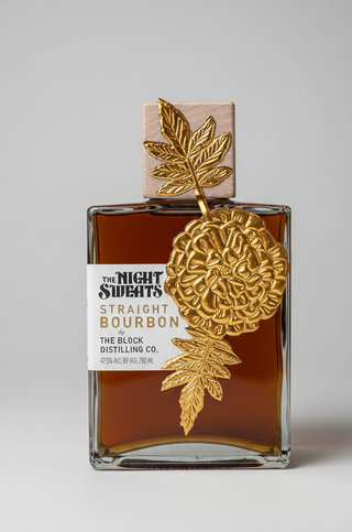 A bottle of Straight Bourbon whiskey from The Night Sweets The Block Distilling Co. with a large gold decorative flower and leaf ornament hanging from the neck, displayed on a glass shelf in a retail setting.